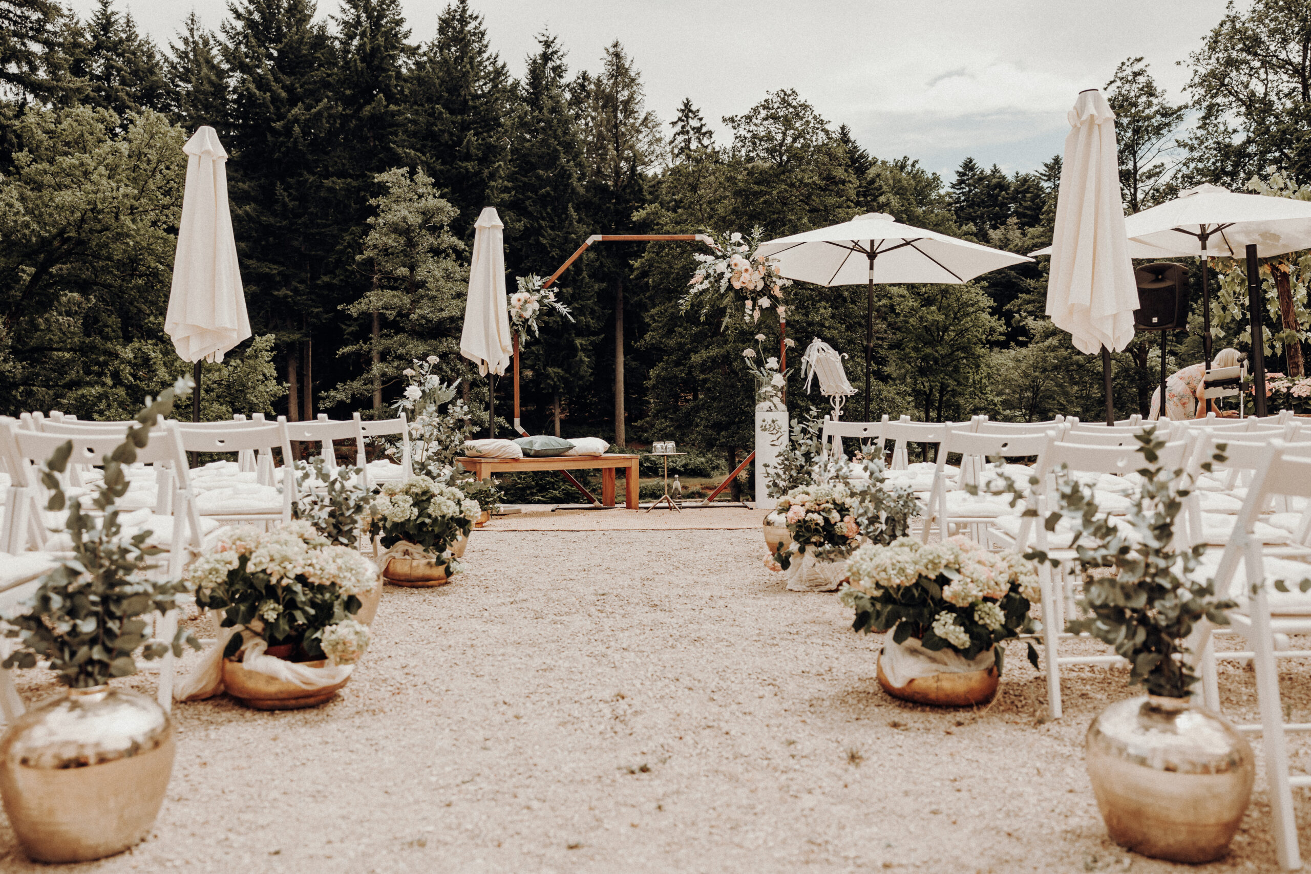 Hochzeitszeremonie im Freien mit weißen Stühlen, Blumenarrangements und Sonnenschirmen unter Bäumen - ein elegantes Dekokonzept, perfekt für Ihre Hochzeitsplanung in Luxemburg.