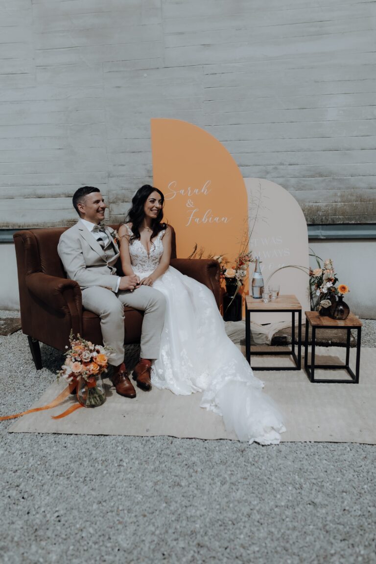 Braut und Bräutigam sitzen bei ihrer saarländischen Hochzeit auf einer braunen Couch, umgeben von eleganter Blumendeko und einem personalisierten Schild, und schaffen so ein atemberaubendes Dekokonzept.