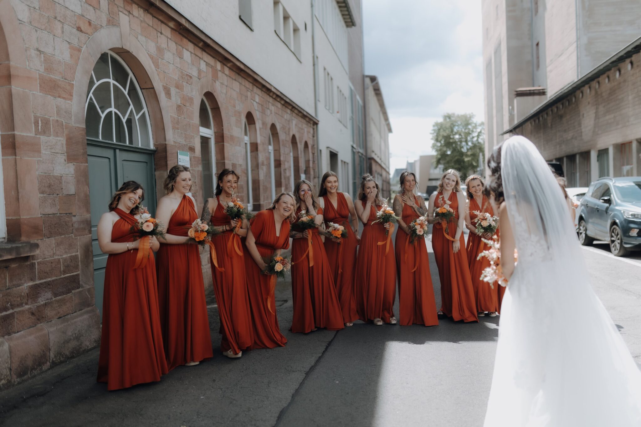 Eine Braut in Weiß steht Brautjungfern in passenden orangefarbenen Kleidern gegenüber, die alle lächelnd im Freien auf einer mit eleganter Blumendeko geschmückten Straße stehen.