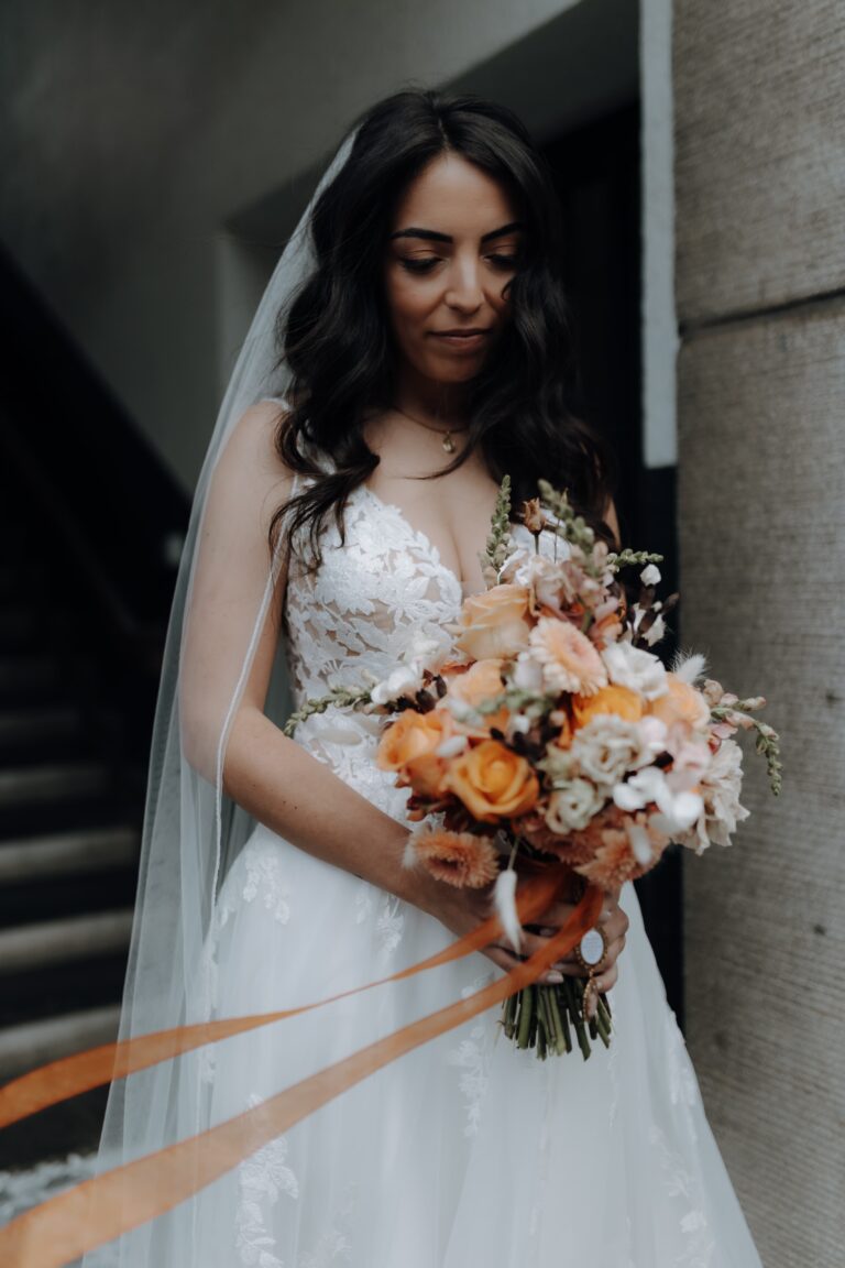 Braut in weißem Spitzenkleid mit einem Strauß orangefarbener und rosafarbener Blumen in der Hand, stehend in der Nähe einer Treppe auf einer schön dekorierten Veranstaltung im Saarland.