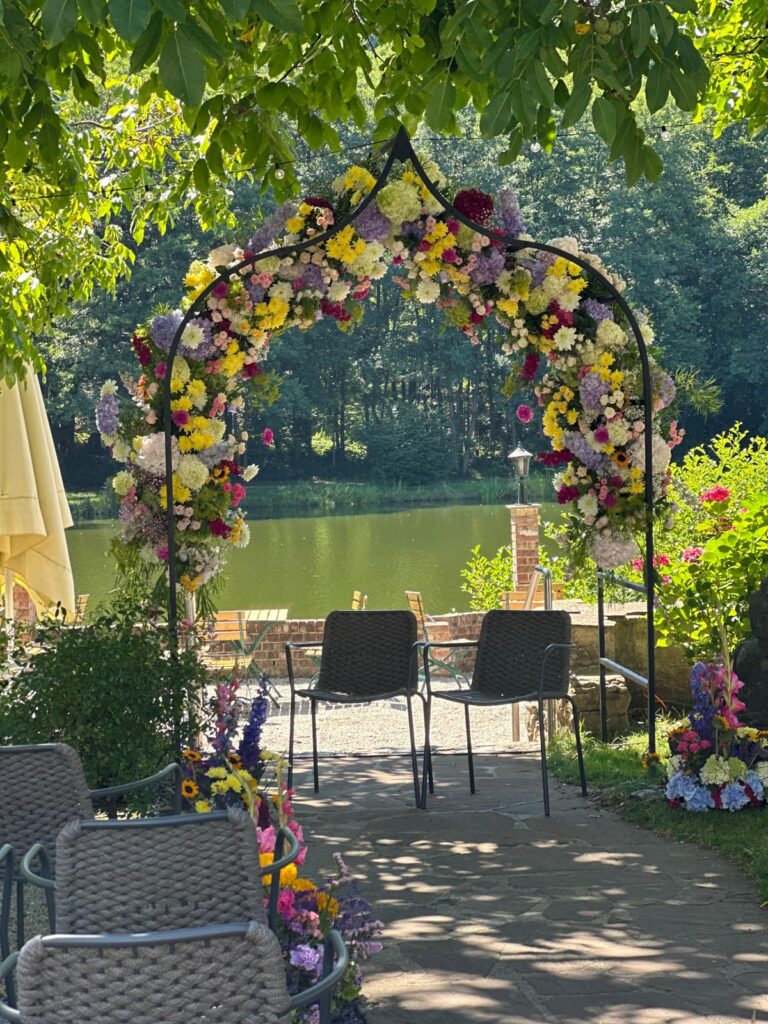 Ein floraler Torbogen umrahmt zwei Stühle mit Blick auf einen See in Luxemburg, umgeben von Grün und Sonnenlicht - perfekt für unvergessliche Veranstaltungen mit einem maßgeschneiderten Dekokonzept.