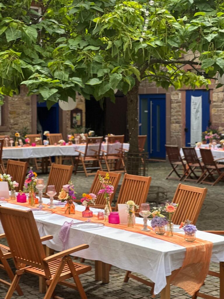Tische im Freien mit Holzstühlen, dekoriert mit Blumen und Kerzen unter belaubten Bäumen in einem luxemburgischen Innenhof - perfekt für stilvolle Veranstaltungen und ein einzigartiges Dekokonzept.