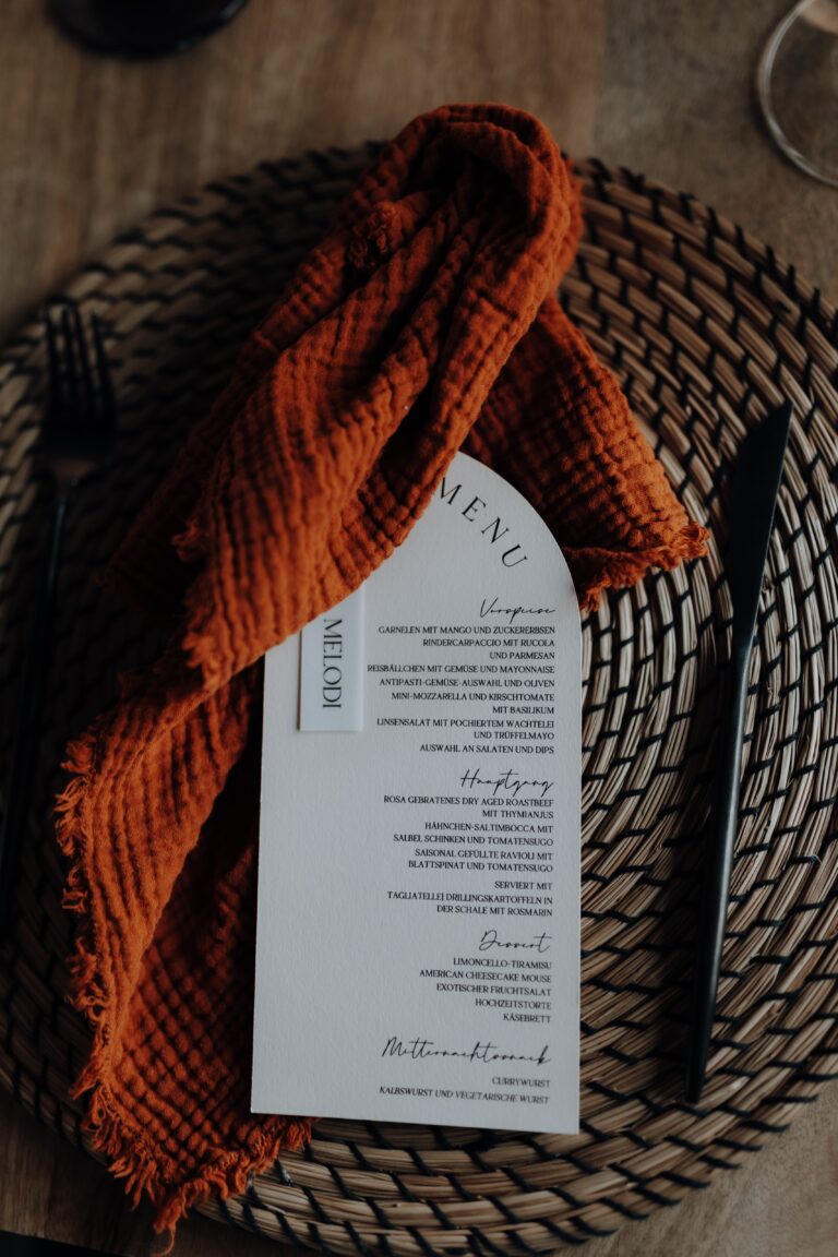 Eine strukturierte orangefarbene Serviette und eine gedruckte Speisekarte liegen auf einem gewebten Tischset, daneben liegt schwarzes Besteck, das ein stilvolles, vom Saarland inspiriertes Dekokonzept widerspiegelt.