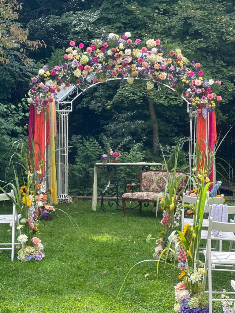 Ein floraler Hochzeitsbogen mit farbenfrohen Blumen und Stühlen in einem üppig grünen Garten ist das perfekte Dekokonzept für unvergessliche Events im Saarland.