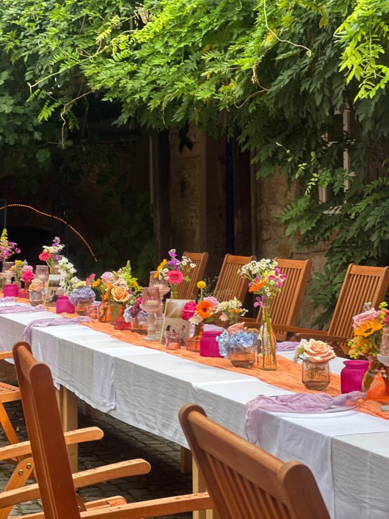 Ein langer Tisch im Freien mit Holzstühlen, leuchtende Blumendeko in farbenfrohen Vasen und festliche Dekorationen in Orange und Pink bilden das perfekte Dekokonzept für unvergessliche Events.