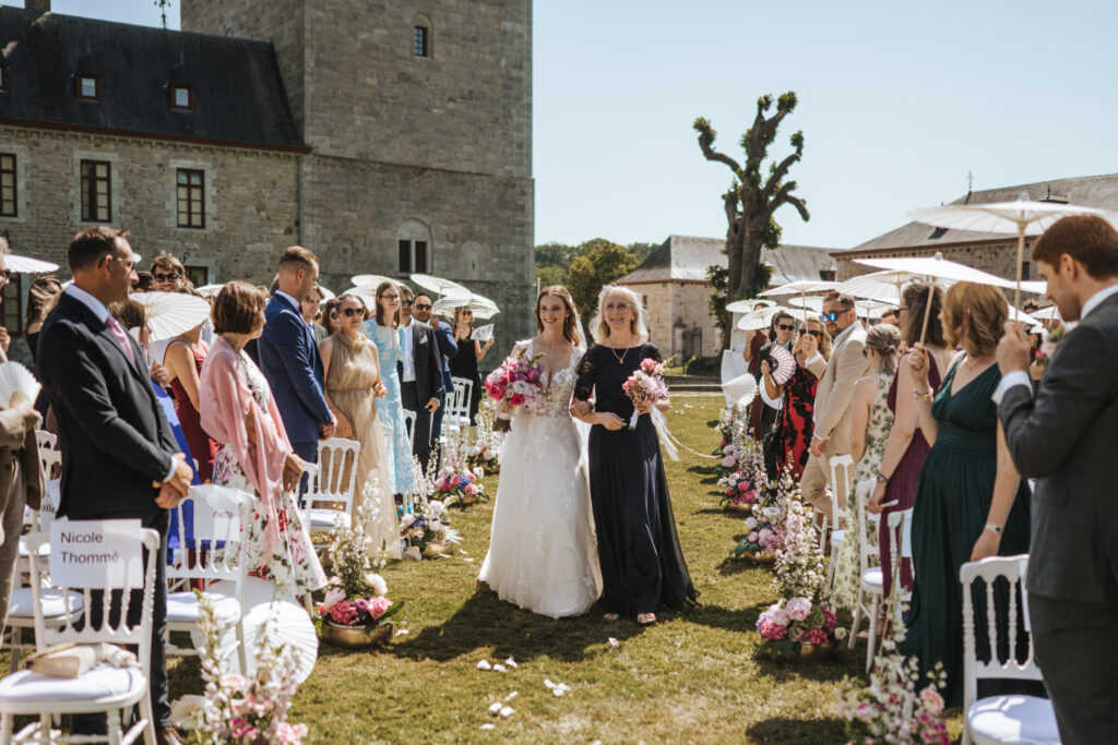 Die Braut im weißen Kleid schreitet mit einem Blumenstrauß in der Hand den mit Blumen gesäumten Gang im Freien entlang, umgeben von einer atemberaubenden Hochzeitsdekoration, während die Gäste zu beiden Seiten zuschauen.