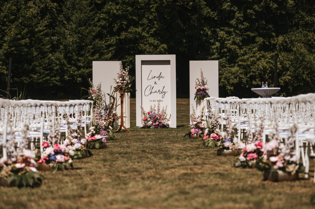 Hochzeitsgang im Freien mit weißen Stühlen, eleganten Blumenschmuck-Arrangements und einem Schild mit der Aufschrift „Linda & Charly, 11.07.2020“.