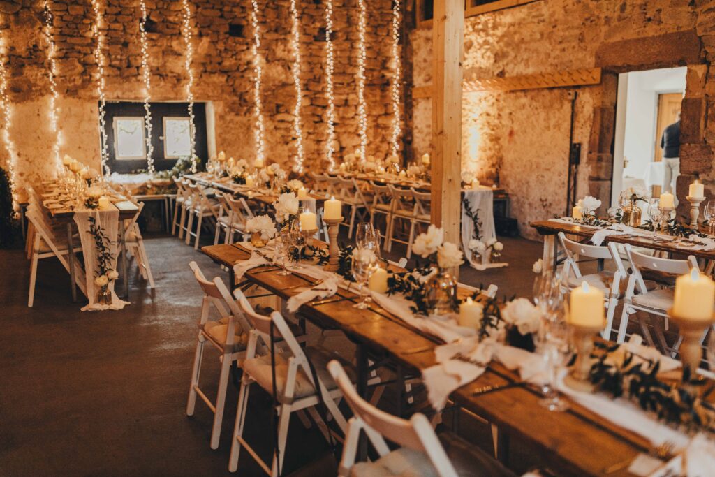 Ein rustikaler Festsaal mit Holztischen, Kerzen, weißen Stühlen, Blumen und Lichterketten – eine bezaubernde Kulisse in der Hochzeitslocation Forsthaus Neuhaus.