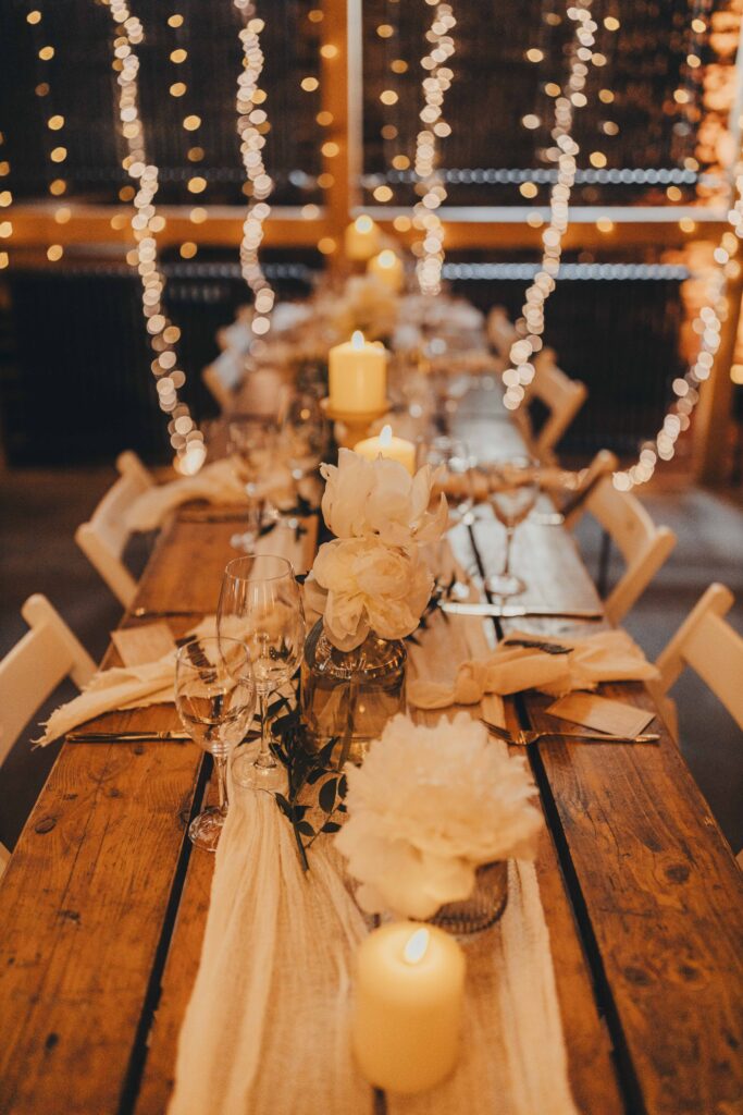 Ein Holztisch, gedeckt für das Abendessen, mit Kerzen, weißen Blumen und Lichterketten im Hintergrund sorgt für eine romantische Atmosphäre – perfekt für eine Feier in der Hochzeitslocation Forsthaus Neuhaus.