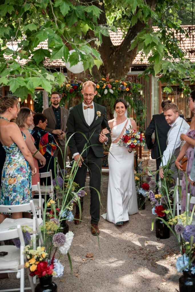 Ein Brautpaar schreitet lächelnd, umgeben von Gästen und farbenprächtigen Blumen, einen Gang im Freien auf der malerischen Hochzeitslocation „Forsthaus Neuhaus“ entlang.