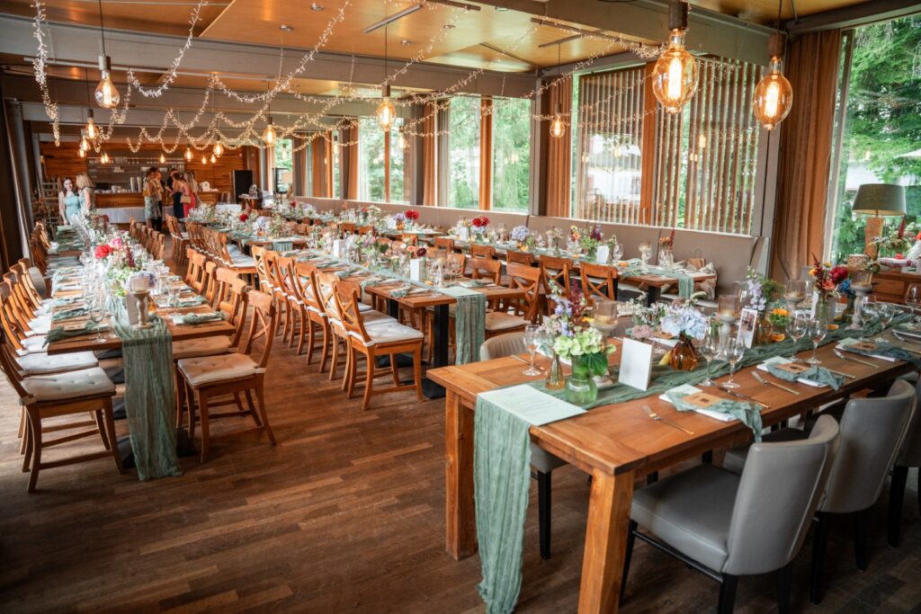 Für eine Veranstaltung in der Hochzeitslocation „Forsthaus Neuhaus“ sind lange Holztische gedeckt, die mit Blumen und grünen Tischläufern unter Lichterketten geschmückt sind.