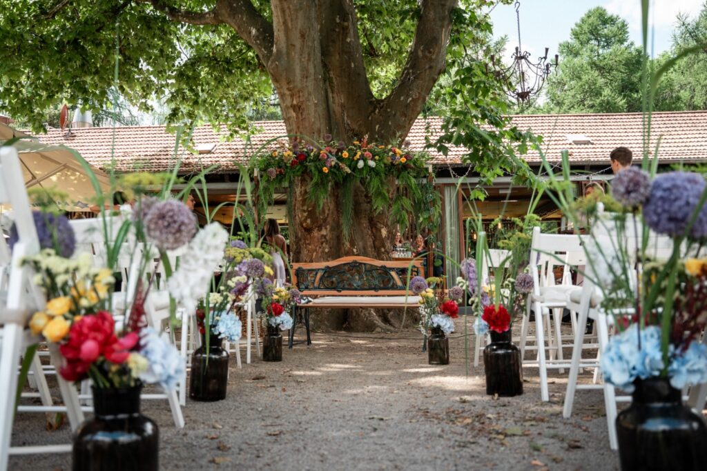 Hochzeitsdekoration im Freien mit Stühlen, bunten Blumen und einer geschmückten Holzbank unter einem großen Baum an der bezaubernden Hochzeitslocation „Forsthaus Neuhaus“.