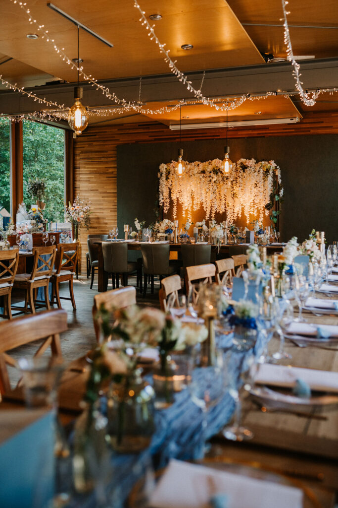 Elegante Hochzeitsfeier in der Hochzeitslocation „Forsthaus Neuhaus“ mit Holztischen, Hängelampen und Blumendekorationen in einem gemütlichen Innenbereich.
