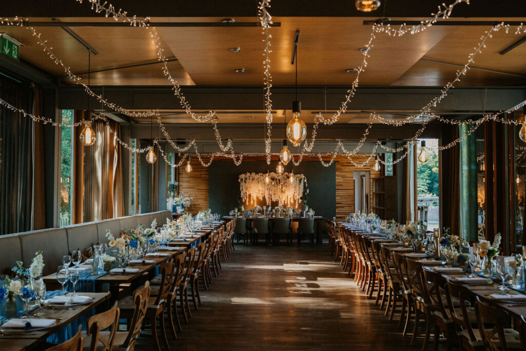 Ein eleganter Festsaal mit langen Tischen, Lichterketten an der Decke und Blumenschmuck auf den Tischen – bereit für eine Veranstaltung: ein exquisites Ambiente, das an die romantische Atmosphäre der Hochzeitslocation Forsthaus Neuhaus erinnert.