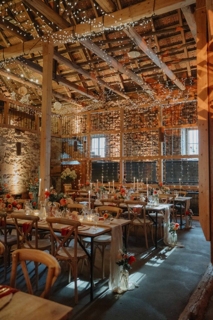 Eine Hochzeit im rustikalen Scheunenstil auf der Hochzeitslocation „Forsthaus Neuhaus“ mit langen Tischen, Stoffvorhängen, Blumen, Kerzen und Lichterketten, die die Holzdecke schmücken.