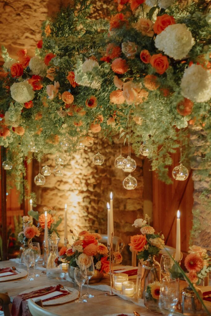 Ein eleganter Tisch mit orange-weißen Blumengestecken, Kerzen und hängenden Glaskugeln in warmem Licht – der das romantische Ambiente der Hochzeitslocation Forsthaus Neuhaus perfekt einfängt.