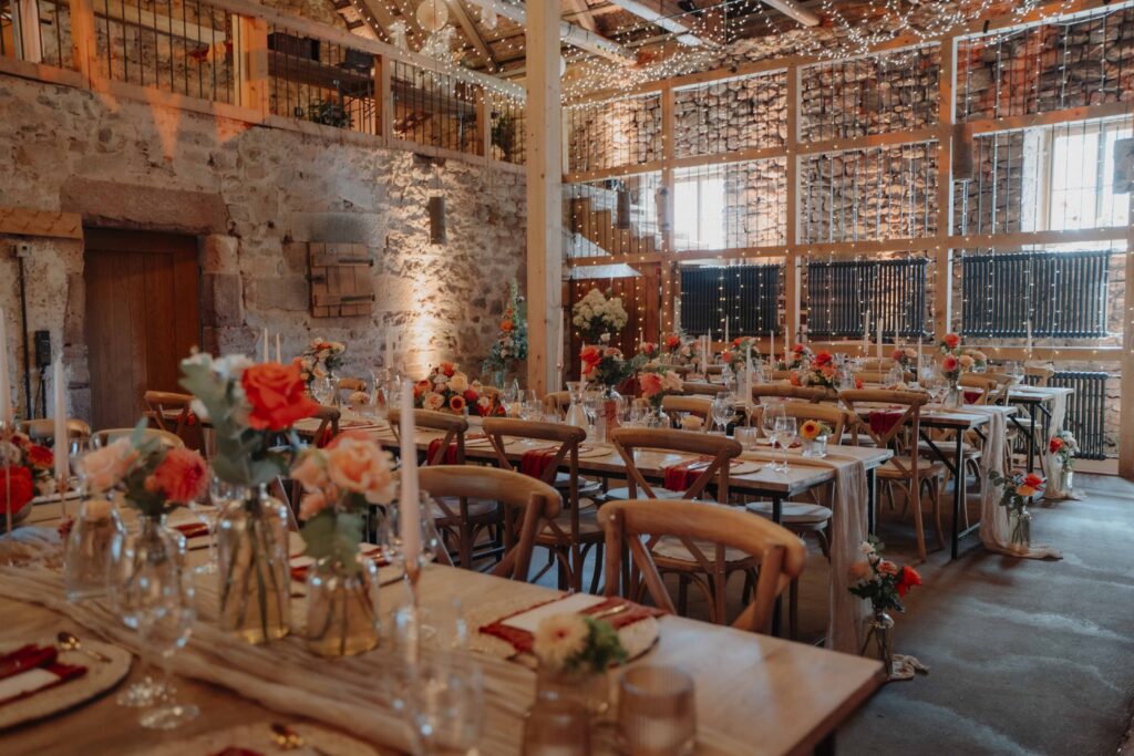 Eine rustikale Hochzeitslocation mit Holztischen, roten Blumen, Kerzen und Lichterketten, die eine Scheune mit Steinwänden schmücken – der perfekte Rahmen für Ihre Feier in der Hochzeitslocation Forsthaus Neuhaus.